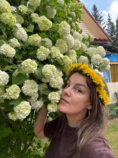 A medium shot shows a young woman with a flower crown of yellow dandelions on her head, posing in front of a bush with numerous white flowers in Malaya Kuskunka, Russia. The woman, facing slightly to the left, has brown hair and is wearing a dark brown t-shirt and a turquoise beaded necklace. Her right hand is extended towards the white flowers. Behind her, a portion of a wooden house with a beige and brown patterned roof is visible, along with green trees and a cloudy sky. The ground in the foreground is covered in green grass.