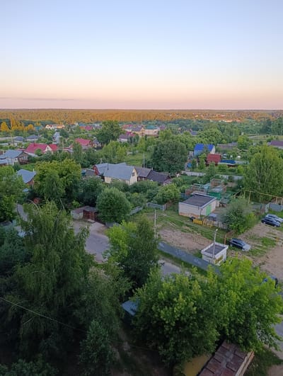 This is an aerial, eye-level view of a rural village in Alexandrov, Russia, bathed in the soft, warm light of either sunrise or sunset. The sky transitions from a pale blue at the top to a soft pink and orange hue near the horizon, suggesting the golden hour.
The scene is a densely packed village with numerous houses, many featuring red or blue roofs, interspersed with lush green trees and vegetation. The houses appear to be predominantly single-family dwellings, with some exhibiting signs of gardening or farming with raised beds and fences visible in several yards. A paved road winds through the village, with at least two cars parked on a gravel area. Several small outbuildings, possibly sheds or garages, are scattered throughout the landscape. In the distance, a dense forest stretches across the horizon. The overall impression is one of a peaceful, everyday life in a rural setting. There is no discernible text in the image.