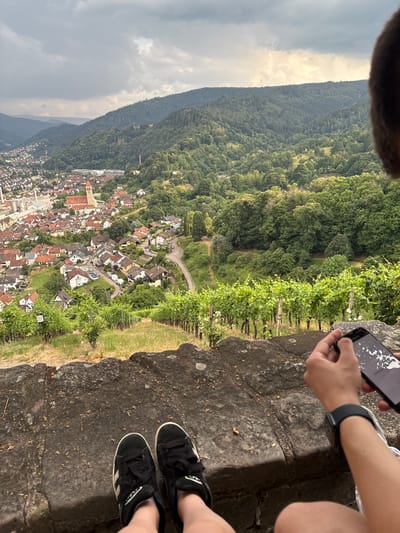The image shows two individuals from the knees down, sitting on a rocky ledge overlooking a town nestled in a valley. One person is holding a smartphone, possibly taking a photo or recording a video of the scene. The town, identified as Gernsbach, Germany, is characterized by its red-tiled roofs and is surrounded by lush green forests and vineyards. The weather appears to be overcast with patches of sunlight, suggesting it could be late afternoon or early morning. The overall mood of the image is one of leisure and appreciation for the scenic vista. There is no visible text in the image.
