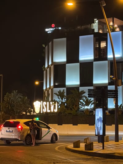 The image captures a nighttime scene on a street in Ibiza, Spain. The primary subjects include a white taxi with its doors open, and a woman who appears to be entering or exiting the vehicle. The taxi has red taillights illuminated and a green and white stripe along its side. A sign with the word "CATS|ST" is illuminated in white, with a decorative element below it.

In the background, a modern building is visible, featuring a facade of alternating dark and light rectangular panels, some of which are illuminated with a cool blue light. A yellow pole with traffic lights or signs is also present, extending across the frame. Various plants and foliage are interspersed throughout the scene, suggesting an outdoor setting, possibly a plaza or street in a well-lit urban area. The time of day is clearly night, indicated by the darkness and the artificial lighting. The overall atmosphere seems to be one of activity, with the taxi and the person interacting with it suggesting movement or travel.