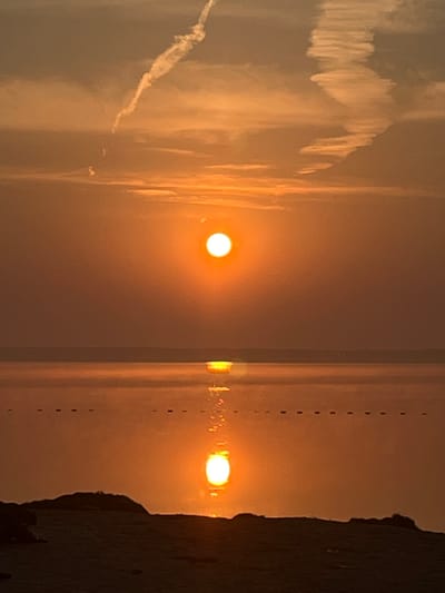 A golden sunset casts a warm glow over a tranquil body of water, with the sun appearing as a bright, luminous orb. The sky is painted with hues of orange and yellow, streaked with wispy clouds and contrails from aircraft. The water reflects the vibrant colors of the sky, creating a mirror-like effect. In the foreground, the dark silhouette of a beach or shoreline is visible, with a few dark objects that might be rocks or debris scattered along its edge. The scene appears peaceful and serene, typical of a sunset over a coastal or lakefront area. The location is identified as Surakovo, Russia.
