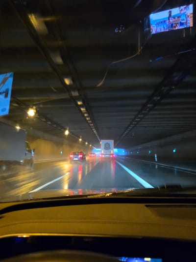 The image depicts the interior of a vehicle driving through a tunnel in Kelsterbach, Germany, during rainy conditions. The tunnel ceiling is dark with rows of yellow lights illuminating the roadway. The wet asphalt reflects the lights and the red taillights of vehicles ahead. A large white truck is visible to the left, and a red car is ahead in the right lane. Further down the tunnel, a white semi-trailer truck with a circular emblem is visible. A small screen is mounted on the right side of the dashboard, displaying a colorful scene. The overall atmosphere is dark and wet, suggesting an evening or nighttime drive.