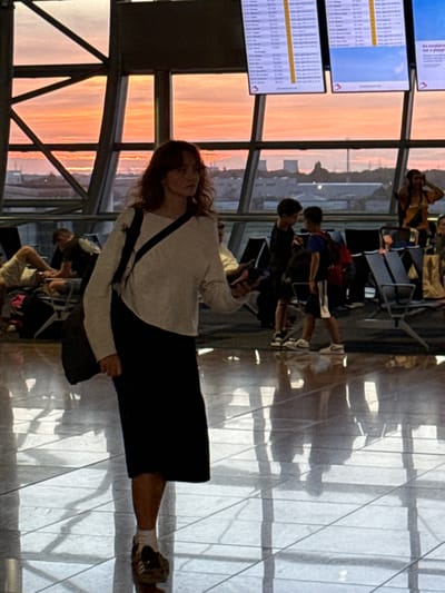 The image depicts a scene inside an airport terminal, likely Brussels Airport in Zaventem, Belgium, given the context of flight information displays. The foreground is dominated by a young woman with reddish-brown hair, wearing a light-colored long-sleeved top and a dark skirt. She is walking towards the viewer and has a dark bag slung across her body. Her feet are clad in white socks and patterned sneakers.
The background reveals a large window with a view of an orange and pink sunset, suggesting it is either dawn or dusk. Through the window, industrial structures and power lines are visible, hinting at a location near an urban or industrial area. Inside the terminal, several other individuals are present. Two young boys are standing in the middle ground, one facing the other. One boy wears a red backpack. In the background, a person is partially visible in an orange top, and further back, passengers are seated in rows of airport chairs. Flight information screens are mounted on the wall above the seating area, displaying text and logos, indicative of an active airport. The floor of the terminal is polished and reflects the surrounding environment and the figures within it. The overall atmosphere is one of travel and waiting.