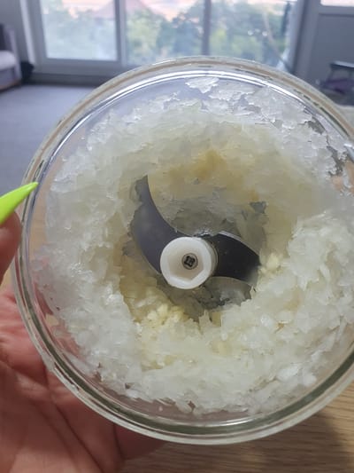 A close-up shot shows the interior of a glass food chopper filled with finely minced white onion. A single silver blade is visible at the center, attached to a white plastic mechanism. The minced onion surrounds the blade and extends to the edges of the glass container. A person's hand, with a neon green fingernail, is visible on the left side of the frame, holding the chopper. The background is out of focus and appears to be an indoor setting with a window showing greenery and a glimpse of distant buildings, possibly indicating a residential area. There is no information in the image to suggest the location is Kheyrabad, Iran.