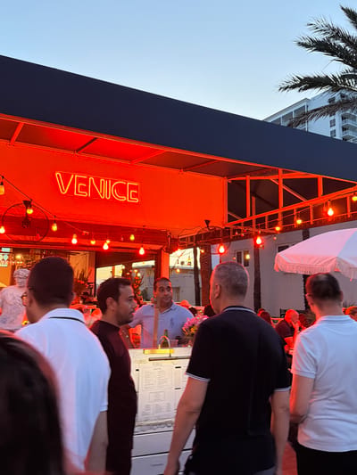 The image depicts an outdoor scene at dusk, likely in the city of Hollywood, United States of America, with a prominent neon sign reading "VENICE" glowing orange. The setting appears to be a restaurant or bar with an open-air setup, indicated by the strung lights and the awning. Several people are visible, engaged in conversations and milling about. In the foreground, several individuals are seen from behind, some wearing casual attire like t-shirts. A white statue, possibly a bust, is partially visible on the left. Behind the counter, a man in a light blue shirt is interacting with someone. The overall atmosphere suggests a lively evening gathering.