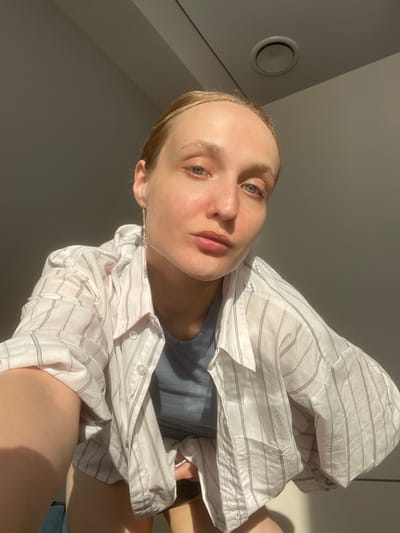 A selfie image features a light-skinned person wearing a gray-blue top and a white shirt with light gray vertical stripes. The subject is positioned at an angle, looking upwards towards the camera. The background consists of a plain wall, with a circular air vent visible in the upper left corner. The image is well-lit, with natural sunlight casting shadows on the wall and the subject's face and clothing. Based on the person's appearance and the style of the image, it is likely a recent photograph taken in an indoor location. It is impossible to determine if the image was taken in Sepych, Russia, or any other specific location based on the visual information alone.