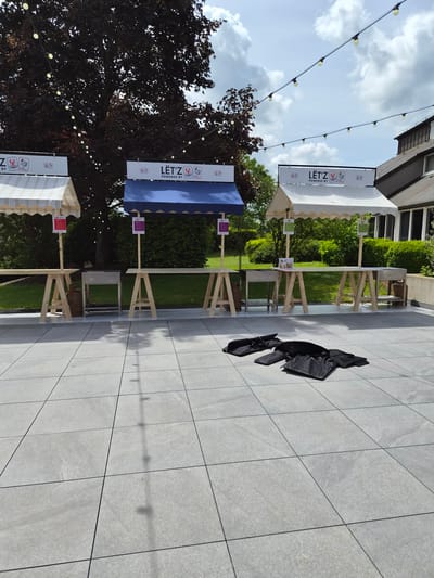 The image shows a partially outdoor event space in Lenningen, Luxembourg. Three wooden tables stand in a row on a gray tiled patio. Each table has a white or blue awning above it, with signs displaying the "Lët'z Powered By" logo. A balled-up black object rests on the tiles near the tables. String lights are strung above. There are buildings and trees in the background.