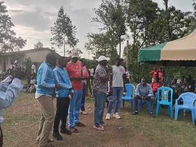 An outdoor gathering is depicted in Chevaywa ward, Kenya, under an overcast sky. In the foreground, from left to right, a person in a light blue long-sleeve shirt operates a black camera, pointed towards a group of men. To their right, two young males in light blue tracksuits stand alongside a man in a red t-shirt and a light-colored baseball cap. Adjacent to them, a man in a patterned short-sleeve shirt and white baseball cap speaks into a handheld object, possibly a microphone, while a man in a grey t-shirt and black baseball cap stands beside him. To the right, a beige and green tent is erected, under which several blue plastic chairs are arranged. One man in a light blue collared shirt is seated on a blue chair, observing the standing speakers. Additional individuals are visible standing and seated under and around the tent. In the background, a single-story building with a light-colored exterior is partially visible, obscured by people and various trees. The ground is a mix of dirt and sparse grass.