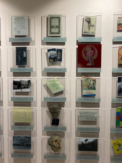 The image shows a wall of framed displays at an exhibition in the Banguntapan district of Yogyakarta, Indonesia.  The display features a variety of artifacts. The artifacts include: 

* A document written in Indonesian.
* A photograph of a group of people in a field.
* A collection of old, handwritten papers. 
* A piece of pottery. 
* A human skull.
* A collection of old photographs and documents. 
* A book called "KANG CRUK" 
* A book called "KHOYSAN DI ATAS BUNIK"
* A book called "Ngaso Sesaat"
* A book called "Ruyung Kembang"
* A collection of photographs.
* A piece of art with a person in a traditional costume. 
* A black and white photo of a person in a traditional costume.
* A photograph of a group of people standing in a field.
* A piece of art with a colorful design. 
* A photo of a person walking with a stick. 
* A piece of art with a design of a person with a stick.
* A photograph of a group of people walking toward a building. 
* A photograph of a person sitting on a bench.
* A piece of art with a design of a group of people.
* A collection of old photos.
* A photograph of a group of people standing together in a field. 
* A piece of art with a colorful design. 

The frames are on a white wall with white borders. The photographs and artworks are arranged in a grid-like pattern on the wall. There are no people in the image.