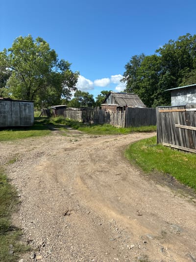 The image depicts a rural scene in the city of Gornye Kljuchi, Russia, characterized by a dirt road winding through overgrown grass and surrounded by weathered wooden structures. The road, composed of gravel and dirt, shows tire tracks, suggesting recent vehicle activity. To the left of the road, a large, metallic-looking shed or building stands, its surface appearing aged and possibly corrugated. Further into the scene, a wooden fence encloses a yard with a small, dilapidated wooden building, its roof made of corrugated material and its walls showing signs of decay. Several other wooden structures and fences are visible in the background, adding to the rustic and somewhat neglected atmosphere. Trees with lush green foliage dominate the background, with a clear blue sky with scattered white clouds above. The overall impression is one of a rural or semi-abandoned area in a natural setting.