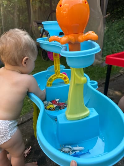 The image shows a close-up of a baby playing with a blue water play table. The baby, with blonde hair, is wearing a diaper and is positioned in the lower part of the frame, facing away from the camera and towards the water table. The water play table is made of blue plastic and has various attachments, including an orange octopus toy and a yellow water wheel. Inside the table, there are several small plastic sea creatures, including a blue whale and a white shark. The scene appears to be outdoors, with a grassy area and some paving stones visible in the background. The lighting suggests it is daytime. The overall impression is one of playful engagement, with the baby focused on the water toys. The image is cropped to emphasize the interaction between the baby and the toy.