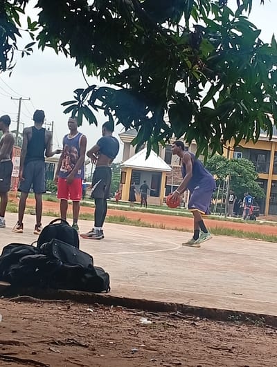 In Ukukwa, Nigeria, a group of young men are engaged in a game of basketball on an outdoor court. The scene is set beneath the dappled shade of trees, with a few buildings visible in the background, one of which is a light yellow, multi-story structure. The sky appears overcast, suggesting a cloudy day.
The central focus is on the basketball players. One individual in the foreground is in mid-air, having just released a basketball. He is wearing a dark purple tank top and matching shorts with yellow trim, along with light green and white basketball shoes. Another player, wearing a blue and white basketball jersey with a graphic print and red shorts, stands watching. To his left, a player in a black tank top and dark blue shorts is also observing the game. Further to the left, another person is partially visible, wearing a black tank top and grey shorts. In the background, other figures are present, including someone wearing a jersey with the number '10' and another with '12', suggesting a broader gathering or multiple games.
A pile of black duffel bags rests on the edge of the court in the foreground, indicating the players' belongings. The basketball court itself is made of concrete with painted lines. The ground surrounding the court is reddish-brown dirt. The overall atmosphere appears to be one of casual sports activity and camaraderie among the young men.