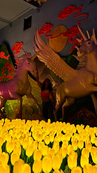 The image depicts an indoor display, possibly within a tourist attraction or museum in Binh Lu, Vietnam. In the foreground is a field of artificial yellow tulips. Behind the tulips, a woman wearing a baseball cap, crop top, and jeans stands between two large, sculpted unicorn statues. The unicorn on the left is more pinkish, while the one on the right has wings and is lighter in color. The statues are set against a painted backdrop featuring red clouds, green hills, and a yellow circle representing the moon. Above this backdrop, there is a grey ceiling with an open vent.