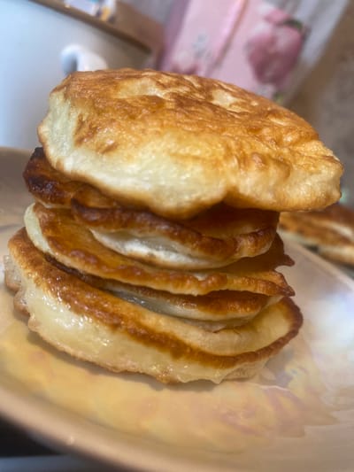 The image is a close-up, shallow depth of field photograph of a stack of golden-brown, fluffy pancakes on a white, iridescent plate. The pancakes are unevenly stacked, with visible air pockets and slightly browned edges. In the background, out of focus, a white mug or cup is visible to the left, and a pink object with floral patterns is to the right. The overall lighting suggests an indoor setting, possibly a kitchen or dining area. The image does not contain any information about the city of Kolomna, Russia.