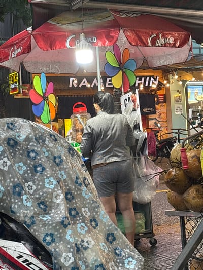 This image captures an outdoor street food scene during the evening in Ngũ Hành Sơn Ward, Vietnam. The ground is wet, suggesting recent rain.
In the foreground, a motorcycle covered by a transparent plastic sheet adorned with blue and white floral patterns is partially visible on the left. On the right, a metal stand holds a cluster of fresh coconuts, some still attached to their stems, with a red handle visible on the stand.
In the mid-ground, an individual, viewed from the back, stands facing a mobile vendor cart. This person has dark hair pulled back and is wearing a horizontally striped grey and white long-sleeved top and matching shorts. They are positioned behind a small, wheeled cart from which several clear plastic bags are hanging. On the cart, a large clear plastic jar with a red lid contains what appear to be fried food items.
The background features a food stall illuminated by a bright LED light bulb suspended from a red and white striped awning branded "Celano." Below the awning, white letters spell "RAMEN HIN" against a dark background, flanked by two large, colorful pinwheels. Various other signs are visible, including a yellow sign with "Chí" and "GOOD HAI," a sign displaying "SHTA'S" and food images, and another sign listing "112 Châu Thị Vĩnh Tế" and "Giờ Mở Cửa 9h-22h." String lights with small bulbs adorn the stall structure. Another motorcycle is partially visible parked further down the street on the right.