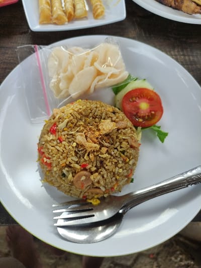 A white plate sits on a wooden table with a serving of fried rice topped with fried onions, a side of fried crackers in a plastic bag, and a tomato and cucumber garnish on the side. Two forks, one on top of the other, are placed on the plate. The plate is at an angle with the camera positioned directly above the plate. The location of the image is a restaurant in Kecamatan Trawas, Indonesia, based on the coordinates provided.