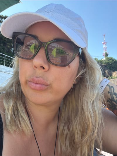A close-up, selfie-style photograph features a woman wearing sunglasses and a white cap. The sunglasses reflect what appear to be green tents or canopies. Her blonde hair is visible around her face and shoulders. A portion of a tower structure is visible in the background against a clear blue sky. A white railing is also present in the background. The woman's right arm, partially visible, has a tattoo and is wearing a white wristwatch. The overall setting appears to be outdoors, potentially near a coast or scenic overlook, given the background elements. The location is identified as Unknown, Ukraine.