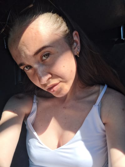 A woman with brown hair is lying down, looking towards the camera. She is wearing a white tank top. Sunlight is casting shadows across her face and body. Her right ear has three piercings. The background is dark and appears to be the interior of a vehicle. The scene suggests a bright, sunny day.