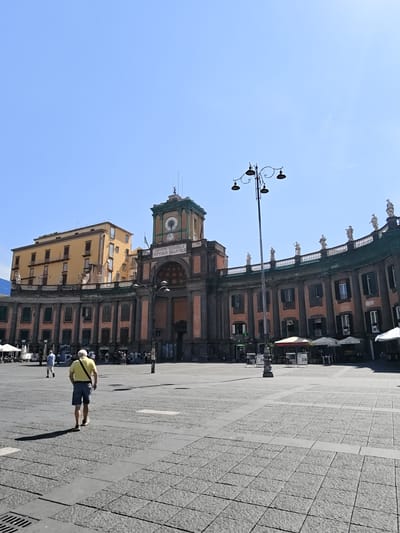 The image shows a view of Piazza del Plebiscito in Naples, Italy, looking towards the Royal Palace. The sun is shining brightly in a clear blue sky. In the foreground, a man wearing a yellow polo shirt and blue shorts walks away from the camera across the large, paved square. The square is made of dark gray and lighter gray stone tiles.
In the midground, a long, curved building with classical architecture forms the background. This building has a dark green roof and is adorned with statues along its upper edge. The facade of the building is a reddish-orange color with dark pillars and windows. To the left of this curved building, a taller, yellow building with multiple stories and balconies stands.
Dominating the center of the image is a large clock tower, which is part of the Royal Palace complex. The clock tower has a greenish-blue roof and a clock face visible. Below the clock tower is a large arched entrance. Several lampposts with multiple lamps are visible throughout the square and in front of the building. Some white umbrellas and outdoor seating areas are set up on the right side of the building, suggesting a cafe or restaurant. A few other people are visible in the distance, walking in the square. The overall scene depicts a bright, sunny day in a historic public square.