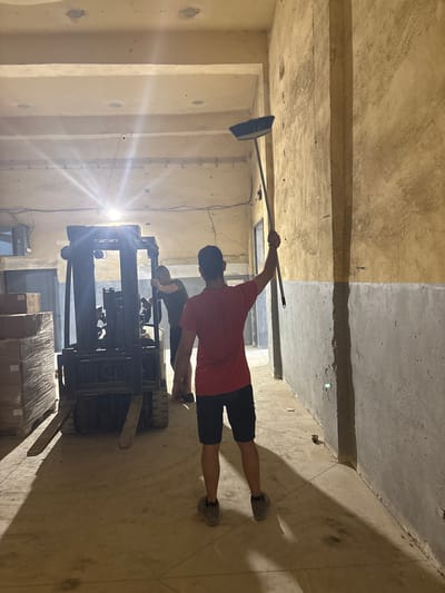 The image shows two people inside a dimly lit, unfinished warehouse or industrial space in Dupnitsa, Bulgaria. One person, wearing a red t-shirt and dark shorts, is positioned in the foreground, performing what appears to be a plank exercise or a similar physical pose, holding a broom with their right arm extended upwards. Their left arm is bent, supporting their body. They are wearing grey athletic shoes. Behind them, another person, wearing a green t-shirt and dark shorts, is hunched over the controls of a black forklift. The forklift has its forks extended and appears to be either parked or in the process of moving.
The setting is characterized by concrete floors and walls, with exposed structural elements and what seems to be unfinished construction. The lighting is harsh, with a strong source of light creating lens flare on the left side of the image, suggesting it might be artificial light from a work lamp or similar fixture. The walls and ceiling show a mix of textures and colors, with some areas appearing lighter and smoother than others, indicating different stages of finishing.
In the background, there are stacks of boxes wrapped in plastic, suggesting that goods are being stored or transported within the space. The overall atmosphere is functional and industrial, possibly a construction site or a storage facility. There is no visible text in the image. The time of day is not explicitly indicated, but the lighting suggests an indoor environment that relies on artificial illumination. The people's postures do not convey any strong emotions, appearing focused on their respective tasks or activities.