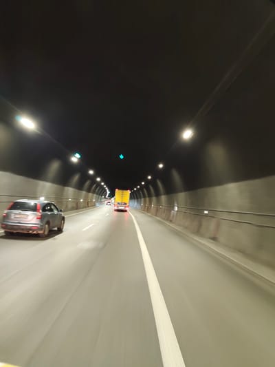 The image captures a forward-looking perspective from within a highway tunnel. The tunnel is illuminated by rows of lights mounted on the ceiling, casting a bright glow on the roadway. The walls of the tunnel appear to be made of concrete. Several vehicles are visible on the road: a silver SUV is in the left lane, and further ahead, a yellow truck with its brake lights illuminated is in the right lane. The road is marked with a white line separating the lanes, and there are some traffic lights visible, showing green signals. The overall impression is one of movement and transit through an enclosed passage. The location is identified as being in the city of Gorno Kamartsi, Bulgaria.