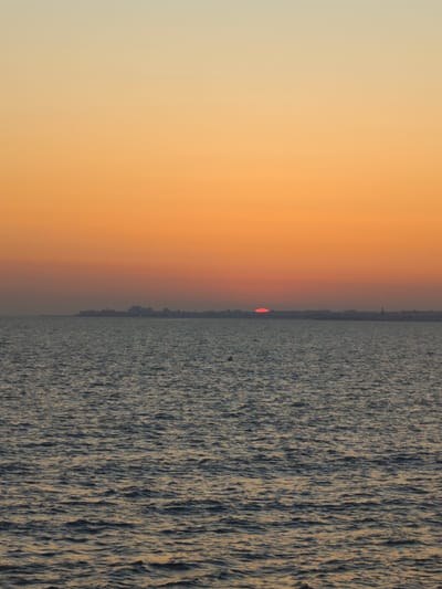 An outdoor, vertical, medium shot captures the tranquil waters of the sea during sunset. The sky transitions from a pale yellow at the top to a warm orange hue near the horizon. A faint, red sun is visible just above a distant landmass, silhouetted against the vibrant sky. The landmass appears to be a city or town, with buildings and structures faintly discernible. The sea itself is a deep, dark blue, with subtle waves reflecting the colors of the sunset. A small, round, yellow buoy is present in the lower left quadrant of the sea. The overall impression is one of a peaceful and serene coastal scene. The location is identified as Costa Oeste, Spain.