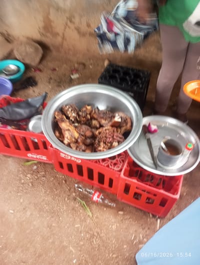 In Dokan Tofa, Nigeria, an outdoor scene displays a gathering around a food preparation area. A large metal bowl, filled with cooked meat, rests on a red plastic crate. Nearby, a silver plate holds a small cup with a spoon, a red onion, and a small bottle with a green cap, possibly containing a sauce or condiment. Another red plastic crate is positioned to the left of the food bowl. In the background, an individual wearing a green shirt and light-colored leggings is visible, along with a patterned cloth and a black plastic container holding bottles. The ground is covered in dirt and scattered debris. A glass bottle is also visible lying on the ground in the foreground. The timestamp "06/16/2026 15:54" is superimposed on the bottom right of the image.