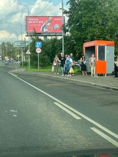 The image shows a bus stop in Novoe Devyatkino, Russia, with several people waiting. The bus stop shelter is orange and grey, and there is a billboard in the background advertising a residential complex. The people are dressed casually and appear to be waiting for a bus. One woman is on her phone, while others are looking in different directions. The scene is set on a paved road with a sidewalk. There are several road signs visible, including a speed limit sign for 40 km/h. Trees and vegetation are present, indicating an outdoor environment. The sky is partly cloudy.