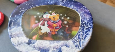 The image shows a close-up, high-angle view of a circular tin with a winter-themed design. The tin has a dark blue background with white snow and bare tree branches. In the center of the tin, through a transparent oval window, are stickers of a snowman and a teddy bear wearing winter clothing. The snowman is white with a purple hat, green scarf with red stars, and a red nose. The teddy bear is orange-yellow with a pink hat, a blue and pink scarf, and a pink coat. It is holding a yellow shovel. The tin contains colorful items that are out of focus. The tin is placed on a gray, textured surface. There are also glimpses of other colorful objects to the left of the tin, one of which appears to be a pink ring. The image is a still life and does not depict any specific location in Cherepovets, Russia.