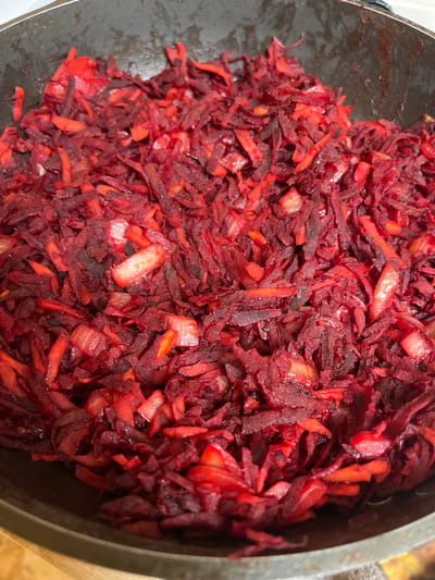 A close-up, overhead shot shows a dark, textured metal pan filled with shredded beets and some lighter-colored ingredients, possibly onions. The beets are a deep, vibrant red, with some pieces appearing darker and more cooked than others. The shredded vegetables are piled high in the pan, filling most of the frame. The lighting is bright, casting some subtle shadows and highlighting the glossy texture of the cooked vegetables. The pan itself appears to be a stovetop pan, with a slightly worn, dark surface. The background is out of focus, but appears to be a wooden surface. The scene is likely a kitchen or cooking environment. No people are visible in the image. The specific location in the city of Vasilyevo, Russia, cannot be determined from the image.