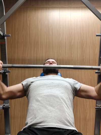 An adult male is observed performing a barbell press exercise on a Smith machine within a gym in the city of Im. Gastello, Russia. The individual, seen from a low-angle perspective, is lying supine on a blue-padded bench, gripping a metal barbell with both hands positioned above his chest. He wears a light grey short-sleeved t-shirt with two faint horizontal stripes on the upper back and dark shorts or pants. The man has a beard and his eyes are closed, suggesting focus during the exercise. The barbell is part of a grey-colored Smith machine structure, with its vertical support frame partially visible on either side of the subject. The background consists of light-toned wooden wall paneling arranged vertically.