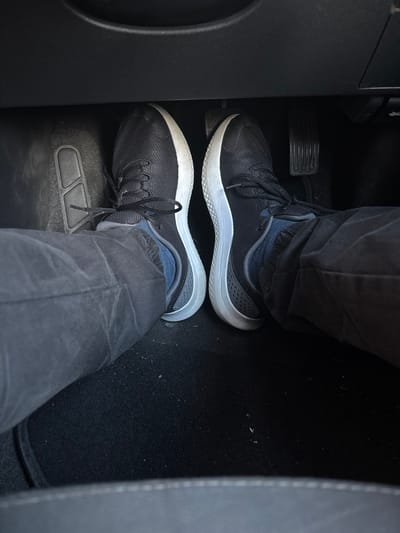 The image shows a first-person perspective of a person's feet wearing black sneakers inside a vehicle, likely a car. The feet are positioned on the floor of the car, with one foot resting on what appears to be a pedal. The pedals are dark, ribbed, and standard for a manual transmission vehicle. The sneakers are black with white soles and dark laces. The person is wearing dark-colored pants. The car's interior is dark and appears to be a standard driver's side footwell. There is no discernible information about the location within the city of Cork, Ireland, or any other geographical markers in the image. The scene is static and offers no indication of action or interaction beyond the placement of the feet.