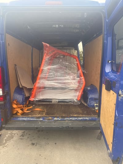 The scene depicts the rear cargo area of a blue commercial van with its double doors open, observed from an exterior vantage point in Logatec, Slovenia. Inside the van, a large, irregularly shaped structure with an orange metal frame is centrally positioned, wrapped entirely in clear plastic film. This object rests on a wooden pallet. To the immediate left of the orange structure, several pieces of brown cardboard are stacked against the van's interior wall. Further left, coiled orange cargo straps with metal hooks are visible on the wooden floor. The interior walls of the cargo area are lined with light-colored plywood paneling. The exterior ground surface beneath the van's open doors is dark grey asphalt or concrete. No individuals are present in the image. The lighting suggests an outdoor, daytime setting.
