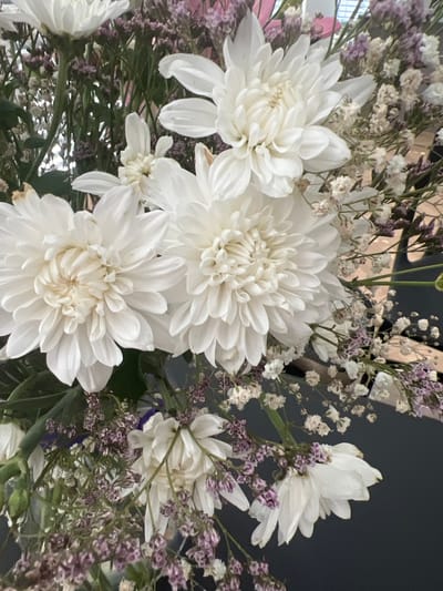 A close-up, overhead shot shows a bouquet of white dahlias and smaller purple and white filler flowers. The bouquet is arranged in a dark grey vase, which is not fully visible. The background is dark and blurred. There are no people in the image. The image is focused on the floral arrangement, highlighting the textures and colors of the flowers. The location of Hof HaCarmel Regional Council, Israel is not depicted or implied in the image.