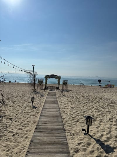 A wooden boardwalk leads across a sandy beach towards a wooden archway decorated with flowers and greenery. The archway is set up on the beach, overlooking the ocean. String lights hang above the boardwalk to the left of the frame, and a palm tree stands to the left of the archway. Several people are visible in the water and on the shore in the background. The sky is clear and blue, with the sun shining brightly. The scene appears to be a wedding or event setup on the beach in Cam Lam, Vietnam.