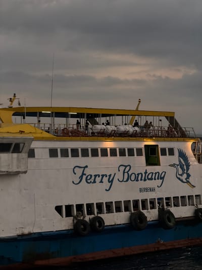 This is a medium shot, eye-level, exterior, static, horizontal, natural light photograph of a ferry boat docked in Melaya, Indonesia. The sky is overcast with dark clouds. The ferry boat is primarily white with a yellow upper deck. The words "Ferry Bontang" are painted in blue cursive on the side of the boat, with "BERBENAH" written below in smaller, blue, capitalized letters. A blue graphic of a bird is depicted to the right of the ferry's name. Several life preservers are visible on the upper deck. People are standing on the upper deck, some appearing to be passengers or crew. The lower portion of the ferry is blue, and it is tied to a dock or other vessel with dark ropes. The water is choppy and dark.
