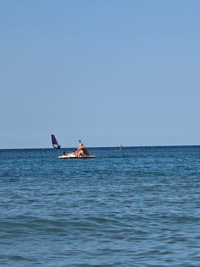 In the city of Ahtopol, Bulgaria, a wide shot captures a calm, blue sea under a clear sky. A pedal boat with a slide is in the middle ground, carrying four individuals. Two people are seated, one appearing to be reclining, and two others are standing. One of the standing individuals is positioned at the rear of the boat, near the slide. Further to the left, a person is engaged in windsurfing, with a purple sail visible. The ocean surface is gently rippled, reflecting the bright daylight.