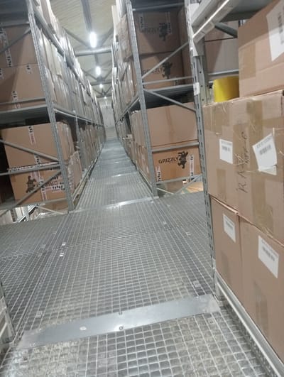 The image shows an interior view of a warehouse with tall shelving units filled with cardboard boxes. The shelves are made of metal, and the flooring appears to be a metal grate. The lighting suggests it is daytime, with artificial lights visible on the ceiling. The boxes are stacked high, some with visible labels that include the brand name "GRIZZLY" and website "www.grizzly.eu". The overall scene depicts a storage facility, likely for manufactured goods. There are no people visible in the image. The location is identified as Aktobe, Kazakhstan.