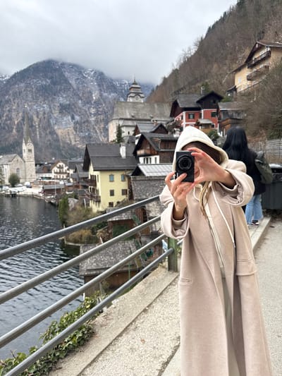 An outdoor scene captures a portion of Hallstatt, Austria, viewed from an elevated pedestrian pathway overlooking Lake Hallstatt. In the foreground, a person faces the camera, holding a black compact digital camera with both hands, obscuring their face. They are wearing a long, light beige hooded coat with visible white drawstrings. To their right and slightly behind, a second individual is partially visible from the back, wearing blue jeans and a dark top, with long dark hair.

The immediate environment includes a gray concrete pathway and a metal safety railing with multiple horizontal bars separating the path from the lake. Green foliage grows along the base of the railing.

The background prominently features the iconic Hallstatt Pfarrkirche (Parish Church of the Assumption) on the left, identifiable by its tall, pointed spire. Further up the steep, tree-covered hillside, the tiered roof of the Beinhaus (Charnel House) or Calvary Church is visible. Numerous traditional alpine-style buildings with gabled roofs, some with balconies, are clustered along the lakefront and ascend the mountainside. The dark water of Lake Hallstatt occupies the lower-left portion of the frame, reflecting the overcast sky. Steep, rocky mountains, partially covered with snow at higher elevations, form the backdrop. The sky is uniformly overcast.