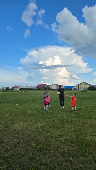 In Bulgakovo, Russia, a man and three young children are on a grassy sports field under a cloudy blue sky. The man, wearing a dark t-shirt and pants, holds a light-colored soccer ball. Two children in pink vests and blue caps stand to his left, and one child in an orange vest stands to his right, reaching out. The field has green grass with scattered patches of dry grass and some white cones. In the background, several houses with red and yellow roofs are visible, along with a large building displaying a sign with Russian text that reads "СВЕТОФОР". A small airplane is visible in the distance in the sky.