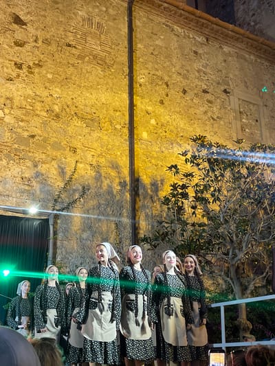 The image shows a group of young women dressed in traditional attire, standing in a line against a stone wall. They are all wearing patterned dresses, aprons, and head coverings, with their hair styled in long braids. Their faces are visible, and some are smiling.
The setting appears to be an outdoor space in Calella, Spain, as suggested by the architectural style of the stone wall and the presence of a tree with dark leaves. The lighting suggests it is evening or night, with artificial lights illuminating the scene. Some of these lights cast a green glow on the subjects.
The women are likely participating in a cultural event or performance, given their synchronized costumes and posed formation. The atmosphere seems festive and engaging. In the background, a part of a white structure is visible, along with some greenery and what looks like a statue. To the right of the frame, a person is holding up a mobile phone, possibly recording the event. No visible text is present in the image.