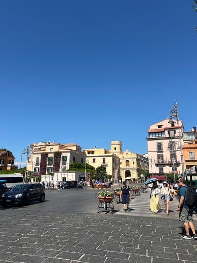 This image captures a bustling street scene in Sorrento, Italy, under a clear blue sky. The foreground is paved with dark cobblestones, and a pedestrian crossing is visible. Several people are crossing the street, including a woman in a long yellow dress and a woman in denim shorts and a white top. Two men are also present in the foreground.
In the middle ground, a dark Volkswagen Up car is seen on the left, and further down the street, a motorcycle with a rider passes by. Several other vehicles and pedestrians are present, suggesting a busy urban environment.
The background features a row of colorful buildings, typical of Italian architecture. One building is adorned with vibrant purple flowers. A prominent yellow church with a bell tower stands out amongst the structures, and several businesses with awnings and signs are visible. The overall impression is a lively daytime scene in a Mediterranean city.