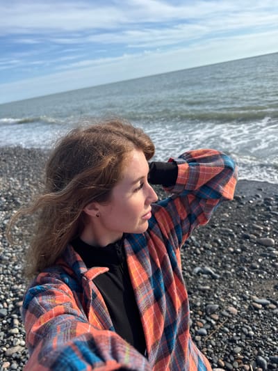 A young woman lies on a pebble beach, facing left. She is wearing a black shirt and an orange and blue plaid flannel. The waves of the sea are at her side. The photograph was taken on the pebble beach in Gulripshi, Abkhazia.