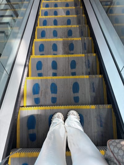 The image shows a top-down view of an escalator with a person standing on it. The escalator steps are gray with yellow edges and blue footprints painted on them. The person is wearing white pants and white sneakers. A patterned bag is partially visible on the top right of the image. The escalator has clear glass sides. The location is not specified as Uttlesford, United Kingdom.