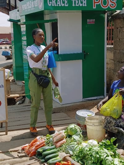 An outdoor market scene is depicted under bright daylight in Communauté rurale de Payar, Senegal. In the foreground, a diverse display of fresh vegetables, including carrots, cucumbers, green beans, cabbage, leafy greens, and tomatoes, is arranged on the ground.
Two females are present. On the left, a young adult female stands, wearing a white t-shirt with green trim, green cargo pants, orange lace-up shoes, and a black fanny pack. She has badges pinned to her shirt and holds a blue item in her left hand and a green item in her right. She is pointing her right index finger towards the left side of the frame, with her mouth slightly open as if speaking. On the right, an adult female is seated on the ground, wearing a blue top and a patterned skirt. Her left hand is near a yellow plastic bag containing produce, and she is looking towards the standing female.
Behind the seated woman, a grey lidded bucket and a white open bucket are visible. In the mid-ground, a green and white kiosk or booth is visible, marked with "PSP P.O.S OUTLET" and "GRACE TECH POS". It is situated partially on a makeshift wooden platform. The background to the left shows a street with parked vehicles and other buildings, while a concrete block wall forms the background to the right.