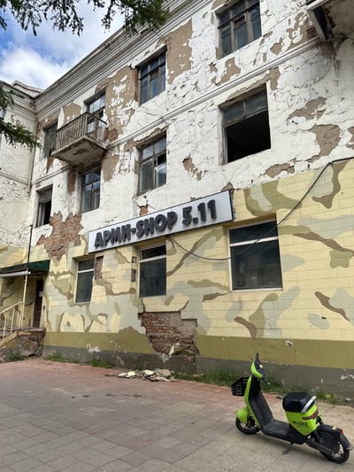 The image depicts a dilapidated building in Ulaanbaatar, Mongolia. The left side of the building has severely peeling white paint, revealing the underlying brick structure. Several windows are visible, some with broken or missing panes. A small balcony is present but in a state of disrepair. The right side of the building features a camouflage-painted exterior, also showing signs of damage and deterioration. A sign reading "APMM-SHOP 5.11" is mounted on the building. An electric scooter, branded "UB BIKE", is parked on the sidewalk in front of the building. The sidewalk is made of red paving stones. The sky is partially visible through tree branches on the left side of the frame.