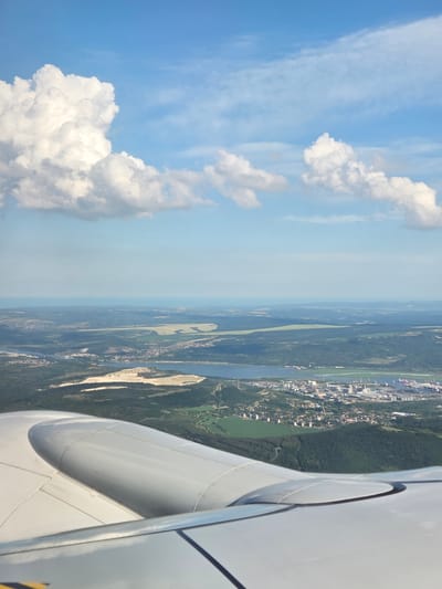 This image, taken from an airplane window, shows a landscape beneath a sky filled with white, fluffy clouds against a bright blue backdrop. The scene is bisected, with the left side dominated by the sky and clouds and the right side revealing the earth below. A portion of the airplane's wing, a large, white, curved object with visible panel lines and a metallic sheen, occupies the foreground on the right.
The landscape below features a body of water, likely a river or a lake, winding through dense green forests. Along the edges of the water, there are signs of human settlement, including buildings and what appear to be industrial facilities or a port, indicated by structures and vessels. Lighter patches in the landscape suggest open fields or potentially sand quarries. The overall impression is of a sunny day, given the clear blue sky and bright illumination of the clouds and land. The image seems to capture a moment of flight, offering a bird's-eye view of a transition zone between natural landscapes and urban or industrial development. No people are visible, and the focus is on the expanse of the sky and the land from an aerial perspective.