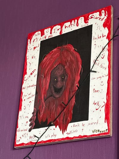 The image shows a close-up of a framed painting hanging on a purple, textured wall. The painting depicts a monstrous figure with red hair and a menacing grin against a black background. The frame is white and has red dripping paint effects, resembling blood. Various phrases and drawings are scrawled on the white frame, including "Hello", "I'll bite you", "I'm good", "No", "Scream", "Not interesting", "I'm not a monster", "It's me", "Hide and seek?", "Don't like?", "Hate", "Let's play", "here?", "friend", "Scared?", "Ho ha ha", "?", "Yes", "Knock Knock", "I'm going to look", "Boo!", "Why?", "- Don't be scared.", "Toothless ?", and a signature that appears to read "UFO Kerry". A string of small, black-wired LED lights is draped across the painting and frame. The location is identified as Radkevshchina, Russia.