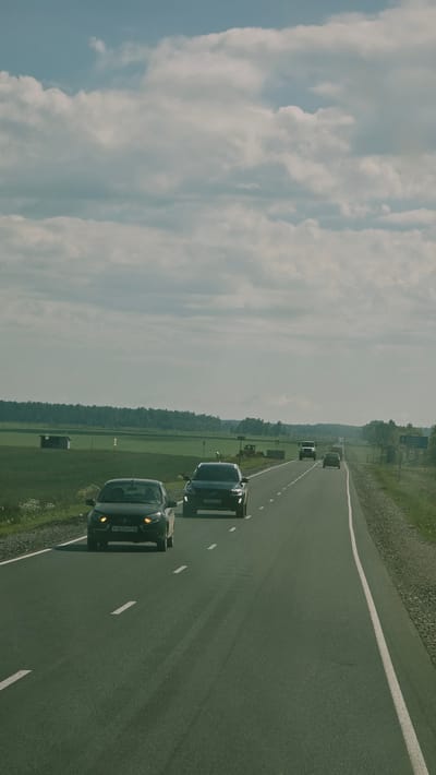 A daylight scene on a rural highway shows several cars driving in the same direction. In the foreground, a dark-colored sedan with license plate T107EM716 is on the left lane. To its right and slightly ahead, a dark SUV with license plate A500XX86 is in the right lane. Further ahead on the right lane, a white truck is visible. The highway is paved and has white dashed lines dividing the lanes. The surrounding environment consists of a grassy field on the left with a small wooden structure and a treeline in the distance. On the right, there is a gravel shoulder and more vegetation. The sky is partly cloudy with large, white clouds. The scene appears to be in a rural area of Актюбинский, Russia, based on the license plates.