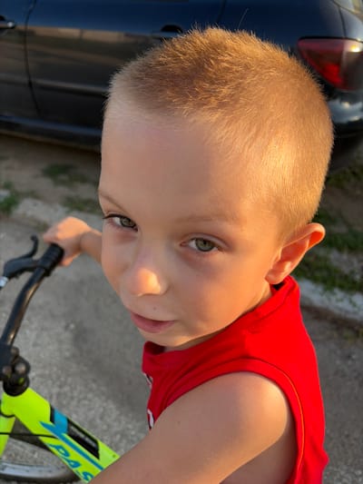 A close-up, eye-level, outdoor photograph shows a young boy with light brown hair and green eyes, looking up and to his left with a slight smile. He is wearing a bright red sleeveless shirt. The boy is holding the handlebars of a neon yellow and blue bicycle, suggesting he is about to ride or has just ridden it. The lighting indicates it is either early morning or late afternoon, with warm sunlight casting a golden glow on his skin and hair. In the background, partially visible to the left, is the dark body of a car, and the ground appears to be asphalt or concrete with some patches of dry grass. The image appears to have been taken on a street or driveway in Dolni Dabnik, Bulgaria.